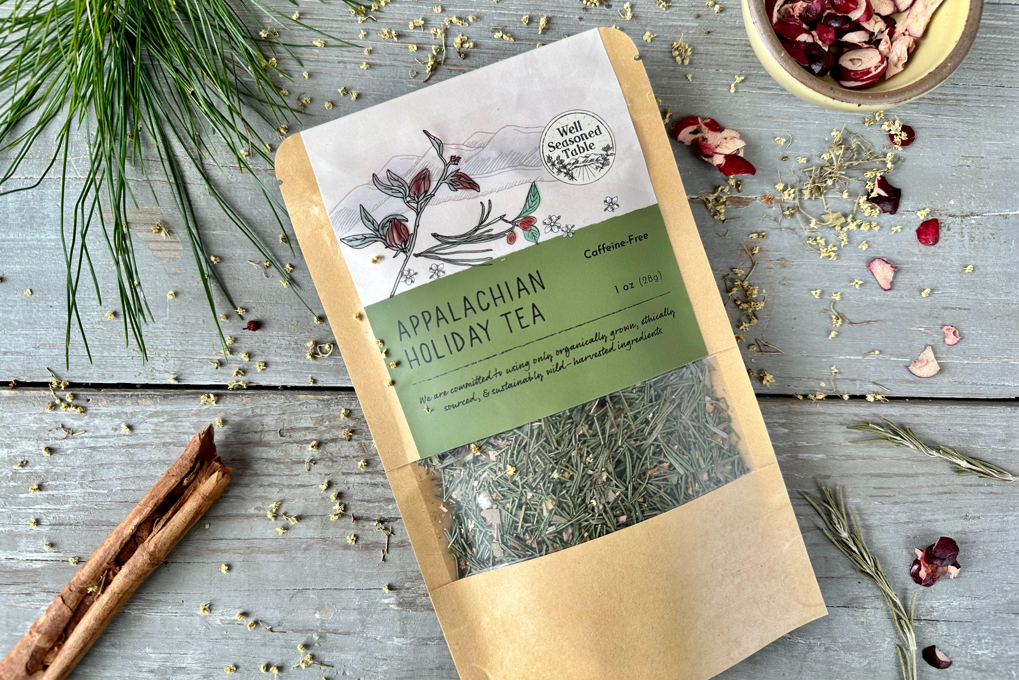 A packet of Appalachian Holiday Tea from Well Seasoned Table on a wooden background with pine needles, dried cranberries, cinnamon, and elder flowers around it.