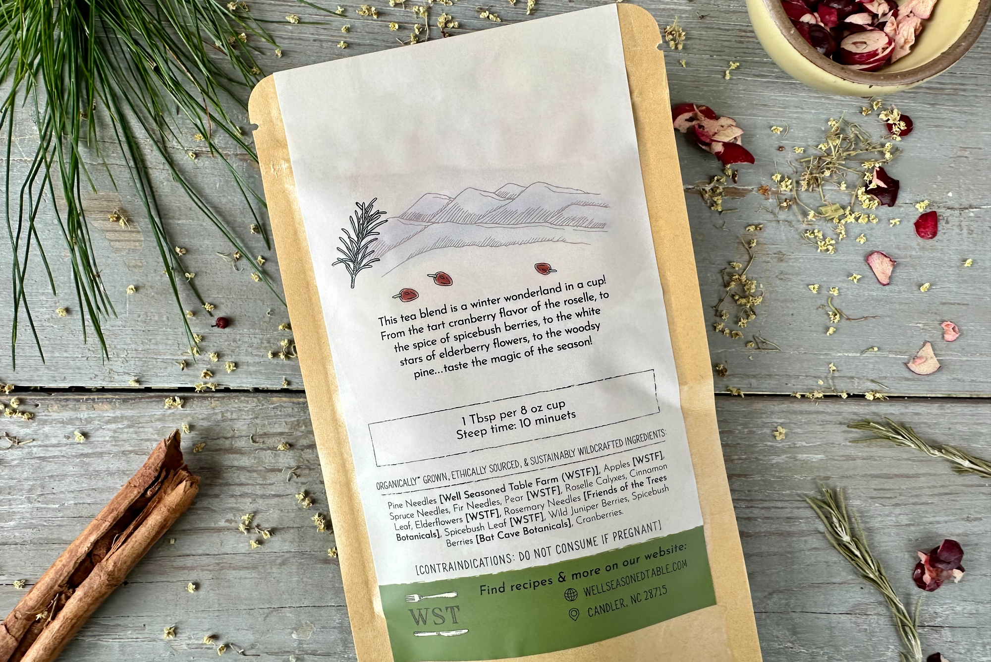 The back of a packet of Appalachian Holiday Tea from Well Seasoned Table on a wooden background with pine needles, dried cranberries, cinnamon, and elder flowers around it.