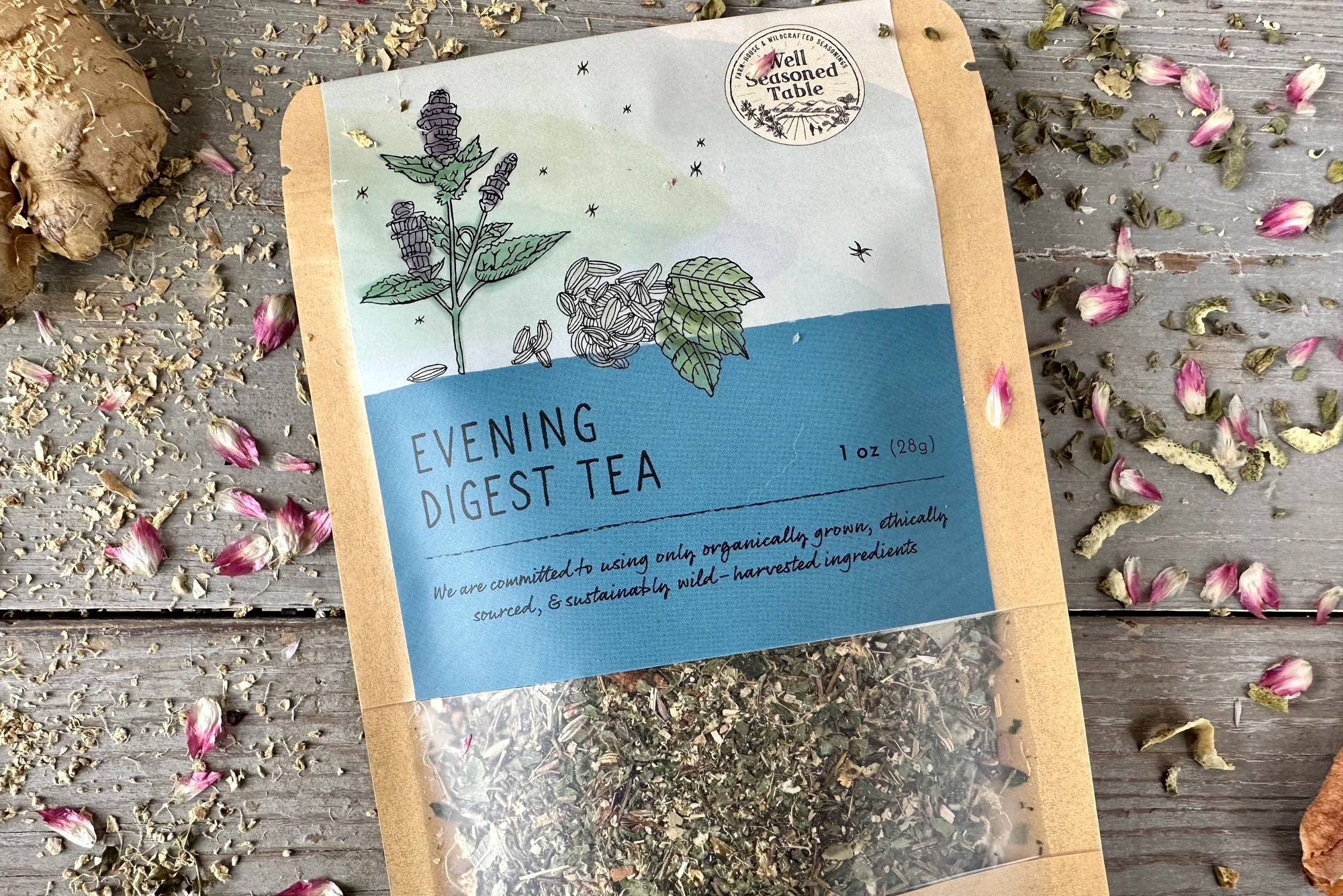 Closeup of a packet of Evening Digest Tea from Well Seasoned Table on a wooden background with ginger, dried flowers, cinnamon, and herbs around it.