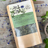A packet of in the garden with tulsi tea from Well Seasoned Table on a wooden background with fresh spearmint and cornflowers around it and a ceramic bowl of tea.