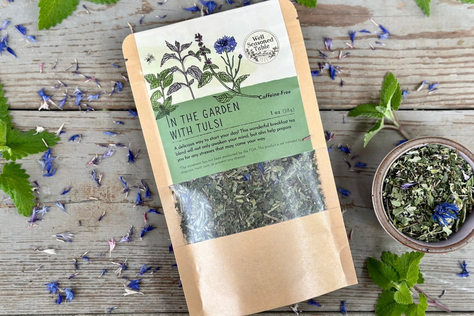 A packet of in the garden with tulsi tea from Well Seasoned Table on a wooden background with fresh spearmint and cornflowers around it and a ceramic bowl of tea.