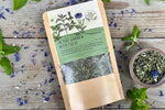 A ceramic bowl of in the garden with tulsi tea from Well Seasoned Table on a wooden background with fresh spearmint and cornflowers around it.