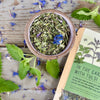 A ceramic bowl of green tea leaves and purple flowers with a packet of In the garden with tulsi tea from Well Seasoned Table on a wooden background with fresh spearmint around it.