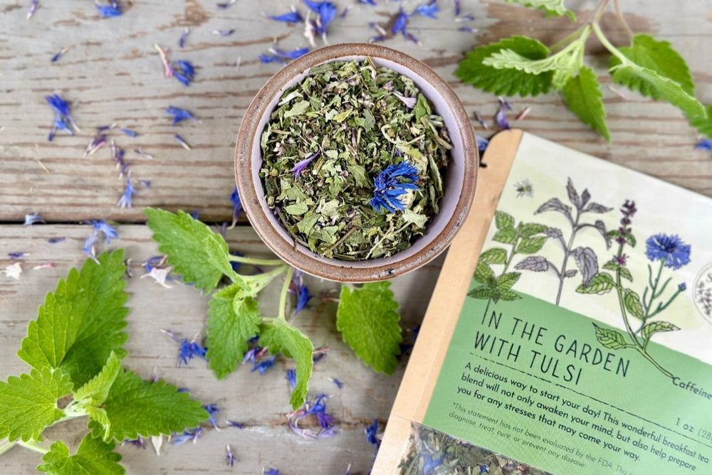 A ceramic bowl of green tea leaves and purple flowers with a packet of In the garden with tulsi tea from Well Seasoned Table on a wooden background with fresh spearmint around it.