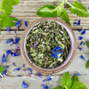 A ceramic bowl of in the garden with tulsi tea from Well Seasoned Table on a wooden background with fresh spearmint and cornflowers around it.