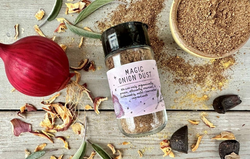 A glass seasoning jar of Magic Onion Dust from Well Seasoned Table on a wooden background with onions, sage, and black garlic around it.