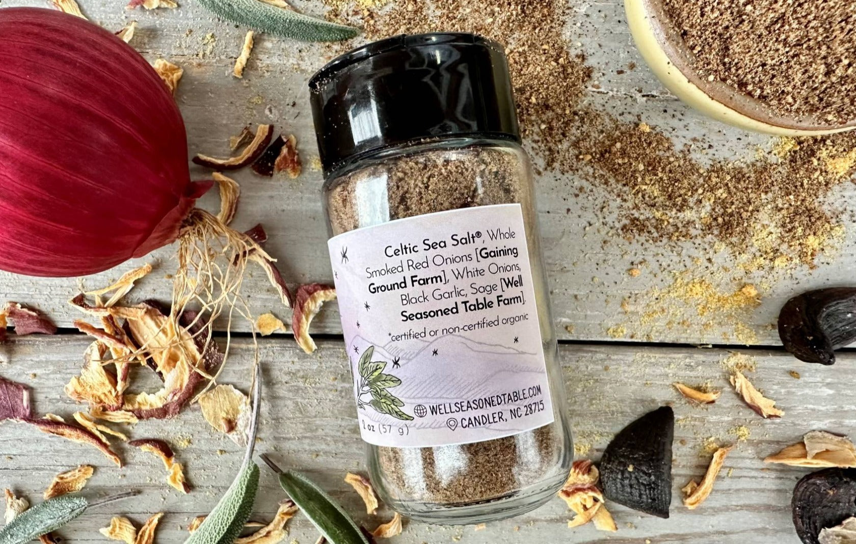 The side of a glass seasoning jar of Magic Onion Dust from Well Seasoned Table on a wooden background with onions, sage, and black garlic around it.