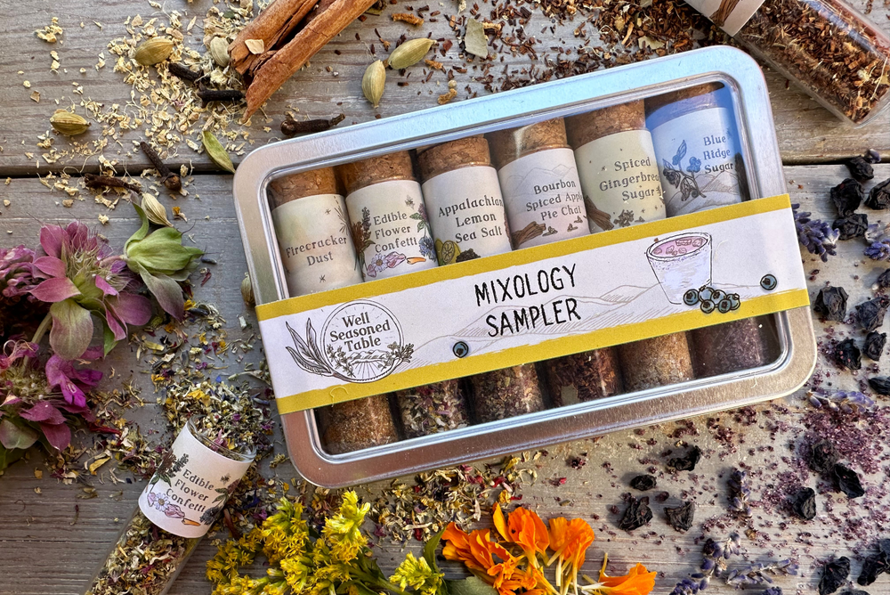 A metal sampler tin with 6 glass vials of infused sugars and organic farm-grown local spice blends and teas surrounded with wildcrafted flowers and dried organic herbs and spices.