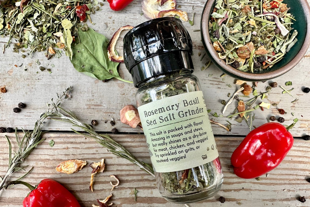 A glass grinder jar of Rosemary Basil Sea Salt from Well Seasoned Table on a wooden background with a ceramic bowl of seasoning, peppers, rosemary, and basil around it.