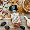 A glass shaker jar of Smoked Fire Seasoning from Well Seasoned Table on a wooden background with a jar of spices and garlic, black garlic, and dried peppers around it.