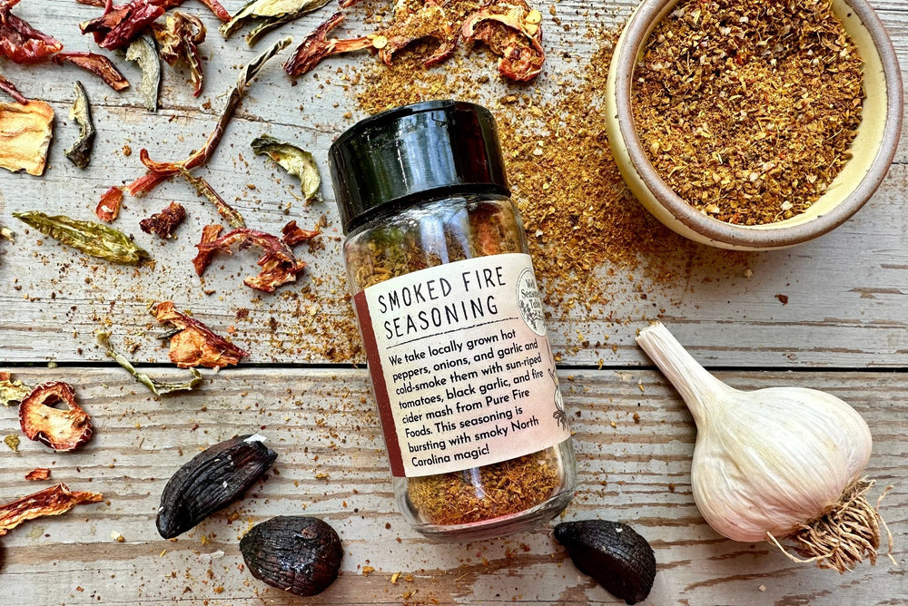 A glass shaker jar of Smoked Fire Seasoning from Well Seasoned Table on a wooden background with a jar of spices and garlic, black garlic, and dried peppers around it.