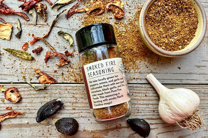 A glass shaker jar of Smoked Fire Seasoning from Well Seasoned Table on a wooden background with a jar of spices and garlic, black garlic, and dried peppers around it.