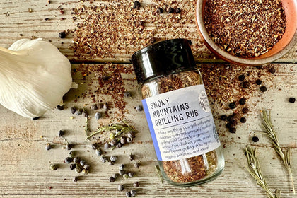 A glass shaker jar of Smoky Mountains Grilling Rub from Well Seasoned Table on a wooden background with a bowl of seasoning, juniper berries, black peppercorns, garlic, and rosemary.
