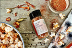 A glass shaker jar of Thai Red Curry Seasoning from Well Seasoned Table on a wooden background with a bowl of seasoning, some seasoned popcorn, and organic dried peppers, ginger, and black peppercorns.