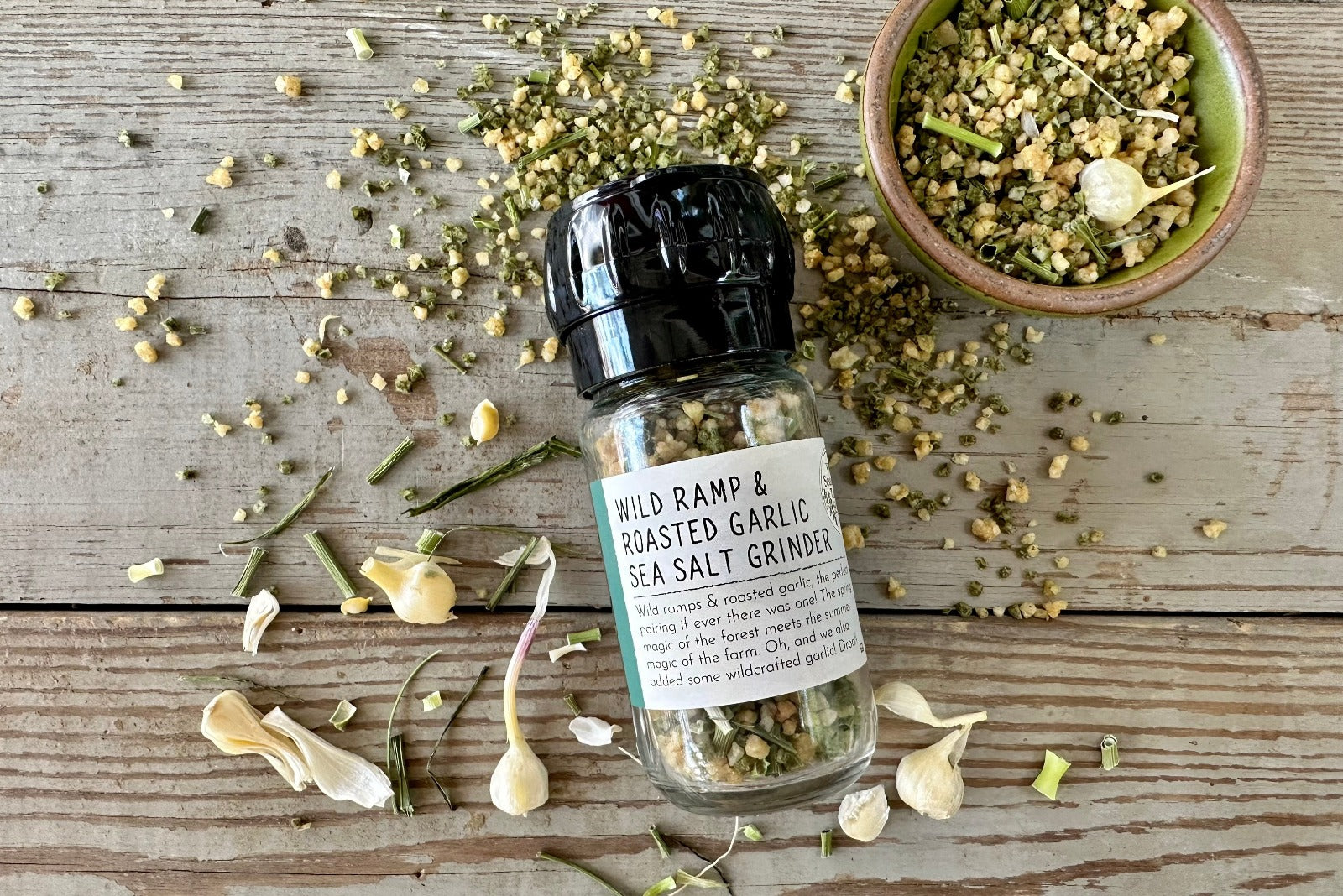 A glass grinder jar of Wild Ramp and Roasted Garlic Sea Salt from Well Seasoned Table on a wooden background with a bowl of product and some dried wild garlic around it.