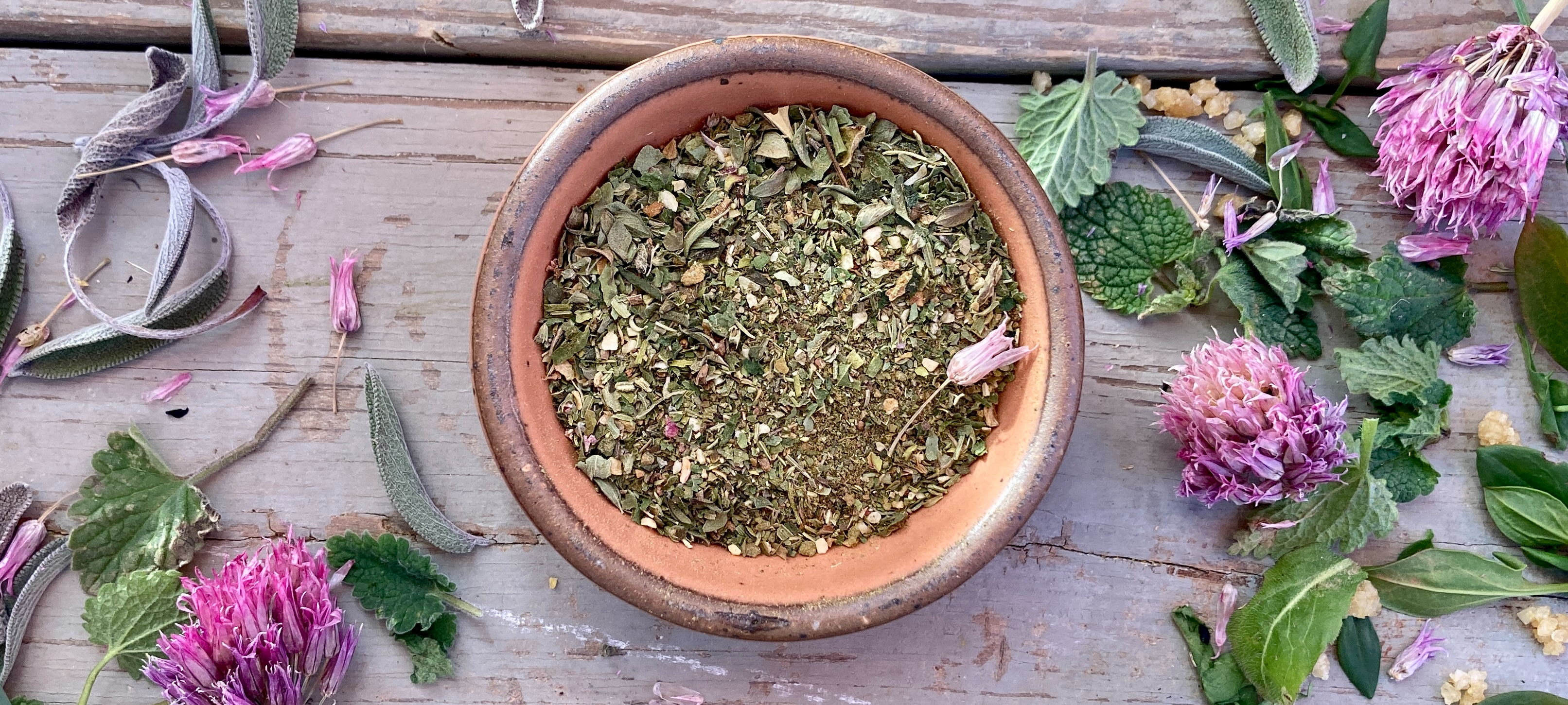 A bowl of Wild Green Goddess Seasoning from Well Seasoned Table on a wooden background with wild greens and chive blossoms around it.