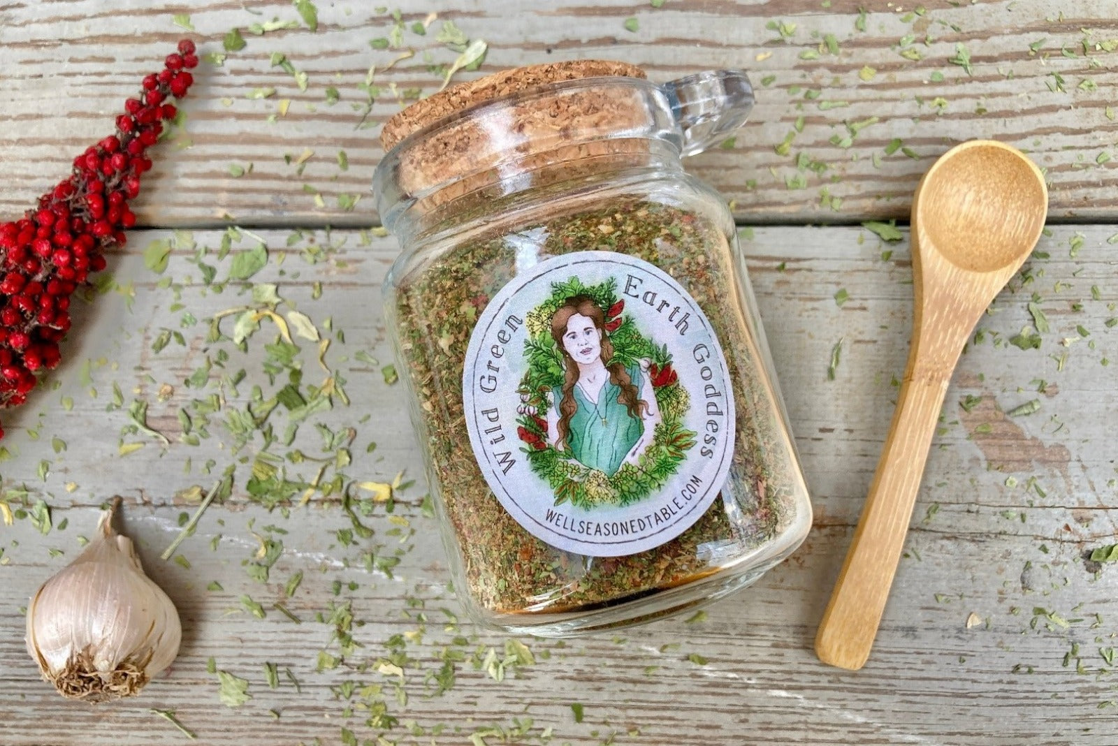 A Glass jar with cork and label and a small wooden spoon on a wooden background with dried herbs, garlic, and sumac around it.