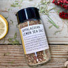 A glass jar of Appalachian Lemon Sea Salt from Well Seasoned Table on a wooden background with dried sumac, lemon, and garlic. 