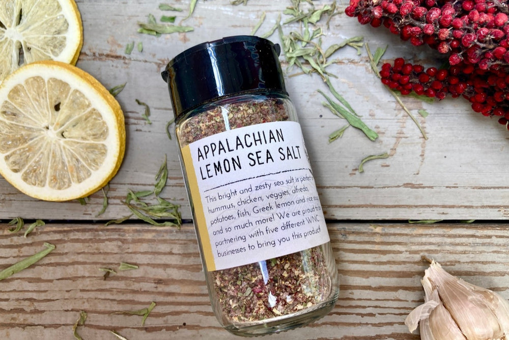 A glass jar of Appalachian Lemon Sea Salt from Well Seasoned Table on a wooden background with dried sumac, lemon, and garlic. 