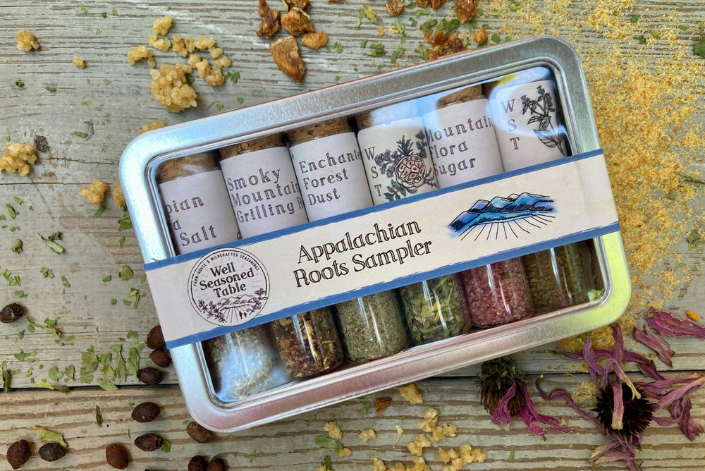 A metal tin of glass vials of organic seasonings from Well Seasoned Table on a wooden background with dried spices and herbs around it. 