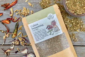 A packet of Cuban Seasoning from Well Seasoned Table on a wooden background with a bowl of seasoning and dried onions and peppers.