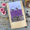 A packet of Dark Chocolate Figgy Pudding Tea from Well Seasoned Table on a wooden background with a cinnamon stick, freeze-dried figs, orange peel, spicebush berries, and dark chocolate chips.