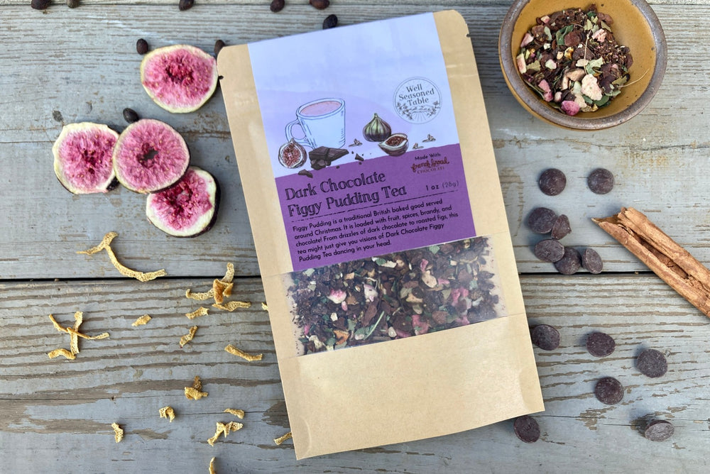 A packet of Dark Chocolate Figgy Pudding Tea from Well Seasoned Table on a wooden background with a cinnamon stick, freeze-dried figs, orange peel, spicebush berries, and dark chocolate chips.