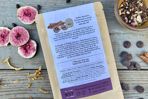 A packet of Dark Chocolate Figgy Pudding Tea from Well Seasoned Table on a wooden background with a cinnamon stick, freeze-dried figs, orange peel, spicebush berries, and dark chocolate chips.