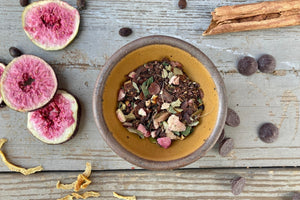 A bowl of Dark Chocolate Figgy Pudding Tea from Well Seasoned Table on a wooden background with a cinnamon stick, freeze-dried figs, orange peel, spicebush berries, and dark chocolate chips.