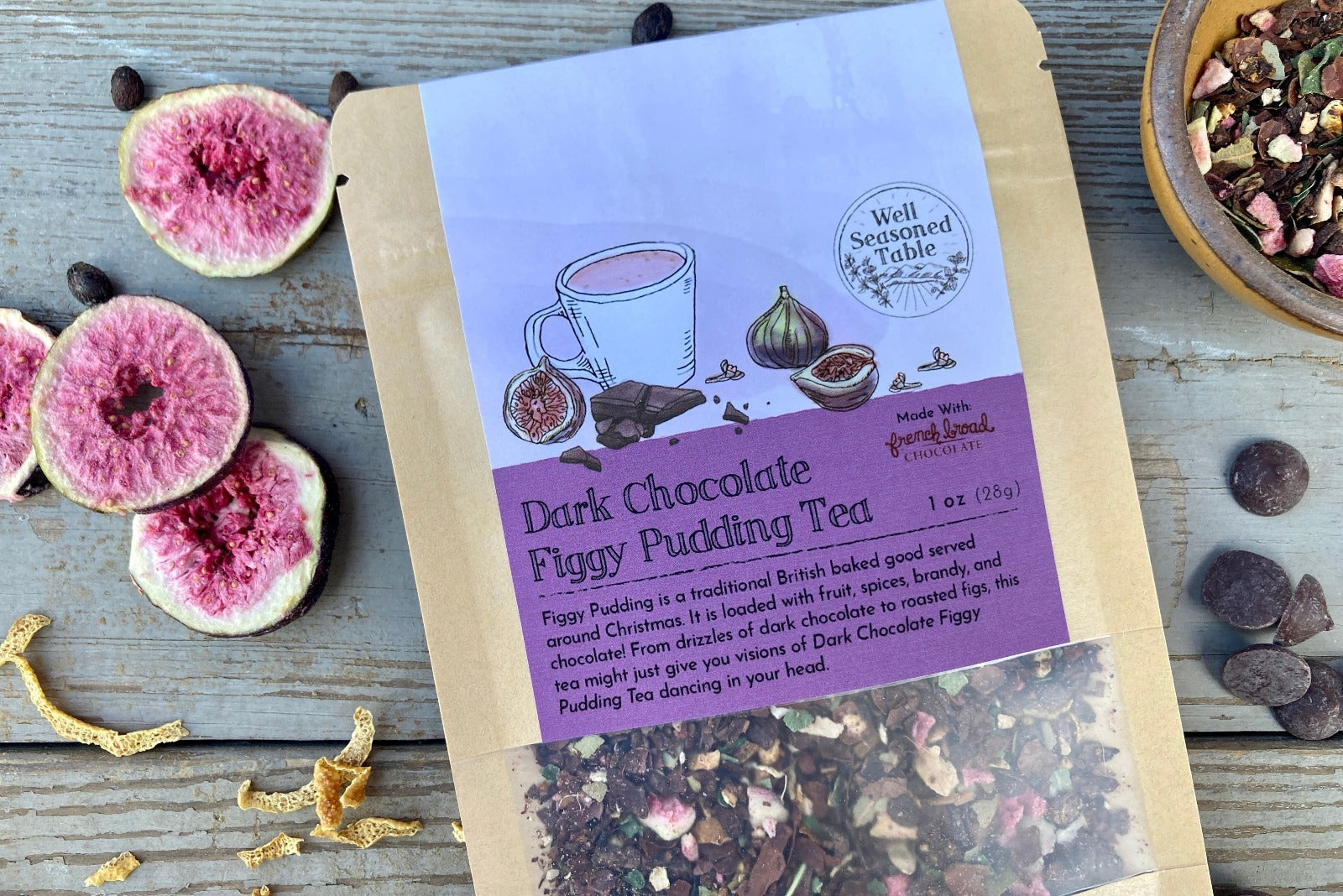 A packet of Dark Chocolate Figgy Pudding Tea from Well Seasoned Table on a wooden background with a cinnamon stick, freeze-dried figs, orange peel, spicebush berries, and dark chocolate chips.