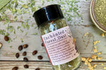 A glass shaker jar of Enchanted Forest dust from Well Seasoned Table, a wild mushroom and herb infused sea salt, on a wooden background with garlic, herbs, ramps, and spicebush berries. 
