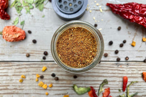A jar of Firecracker Dust from Well Seasoned Table on a wooden background with assorted spices and herbs around it.