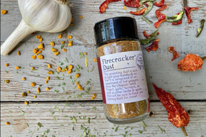 A glass shaker jar of Firecracker dust, an organic spice blend from Well Seasoned Table, on a wooden background with garlic, peppers, and herbs around it. 