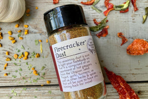 A glass shaker jar of Firecracker dust, an organic spice blend from Well Seasoned Table, on a wooden background with garlic, peppers, and herbs around it.
