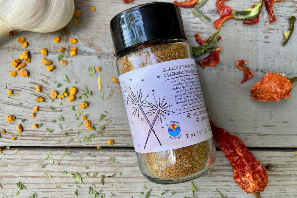 A glass shaker jar of Firecracker dust, an organic spice blend from Well Seasoned Table, on a wooden background with garlic, peppers, and herbs around it.
