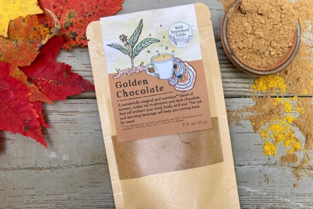 A packet of Golden Chocolate from Well Seasoned Table on a wooden background with a bowl of cocoa powder and a sprinkle of turmeric. 