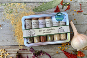 A metal tin of glass vials of organic spice blends from Well Seasoned Table on a wooden background with dried garlic, peppers, wildcrafted ramps, and dried flowers around it.