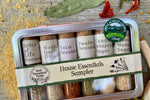 A metal tin of glass vials of organic spice blends from Well Seasoned Table on a wooden background with dried garlic, peppers, wildcrafted ramps, and dried flowers around it.