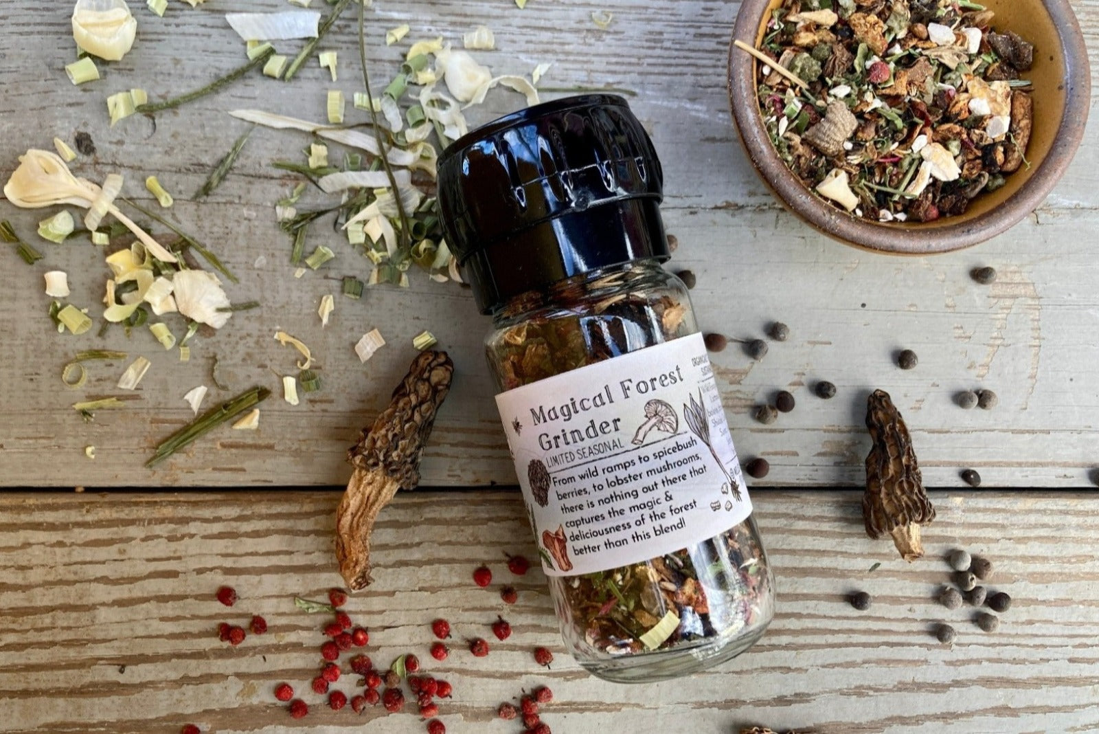 A glass grinder jar of Magical Forest Grinder from Well Seasoned Table on a wooden background with wild onions, dried morels, and sumac berries around it. 