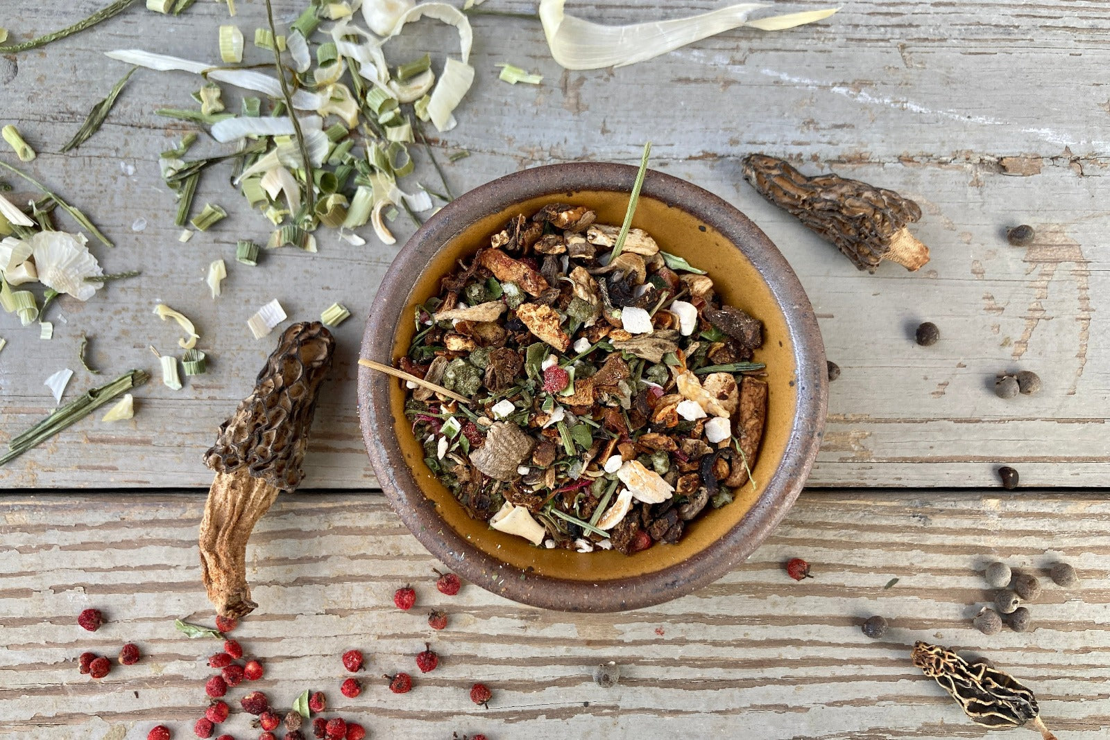 A bowl of Magical Forest Grinder from Well Seasoned Table on a wooden background with wild onions, dried morels, and sumac berries around it. 