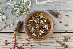A bowl of Magical Forest Grinder from Well Seasoned Table on a wooden background with wild onions, dried morels, and sumac berries around it. 