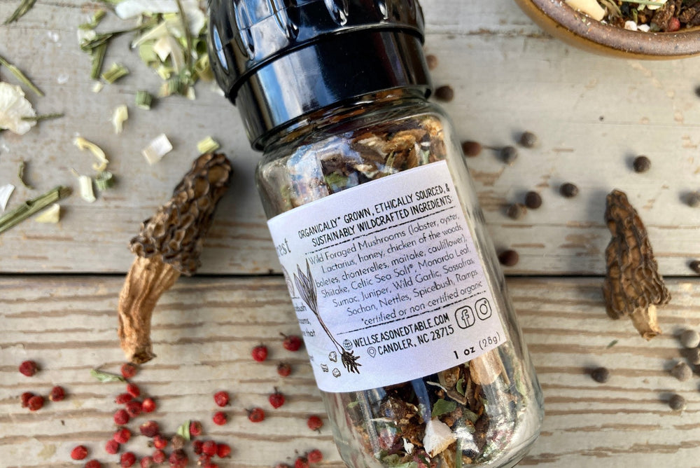 A glass grinder jar of Magical Forest Grinder from Well Seasoned Table on a wooden background with wild onions, dried morels, and sumac berries around it. 