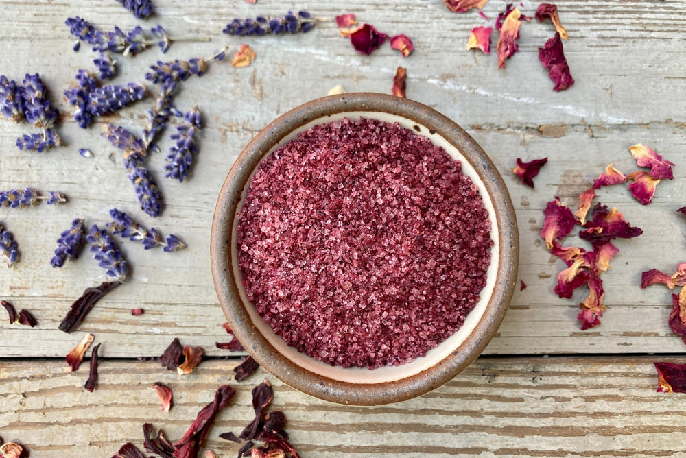 A bowl of Mountain Flora Sugar, an organic infused sugar, from Well Seasoned Table, a spice shop, on a wooden background with hibiscus, lavender, and rose petals around it.