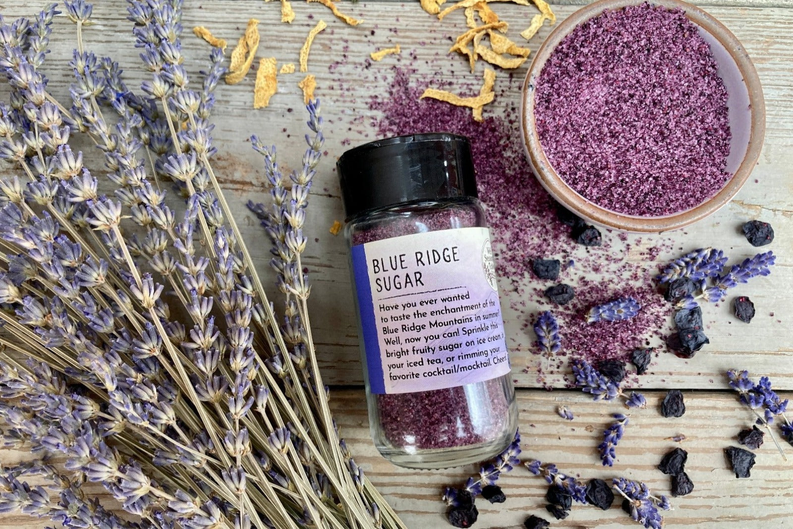 A glass shaker jar of Blue Ridge Sugar from Well Seasoned Table on a wooden background with dried lavendar, lemon, and blueberries around it.