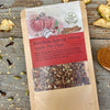 A packet of Bourbon Spiced Apple Pie Chai Tea from Well Seasoned Table on a wooden background with dried apples, spices, and a spoon of cinnamon around it.