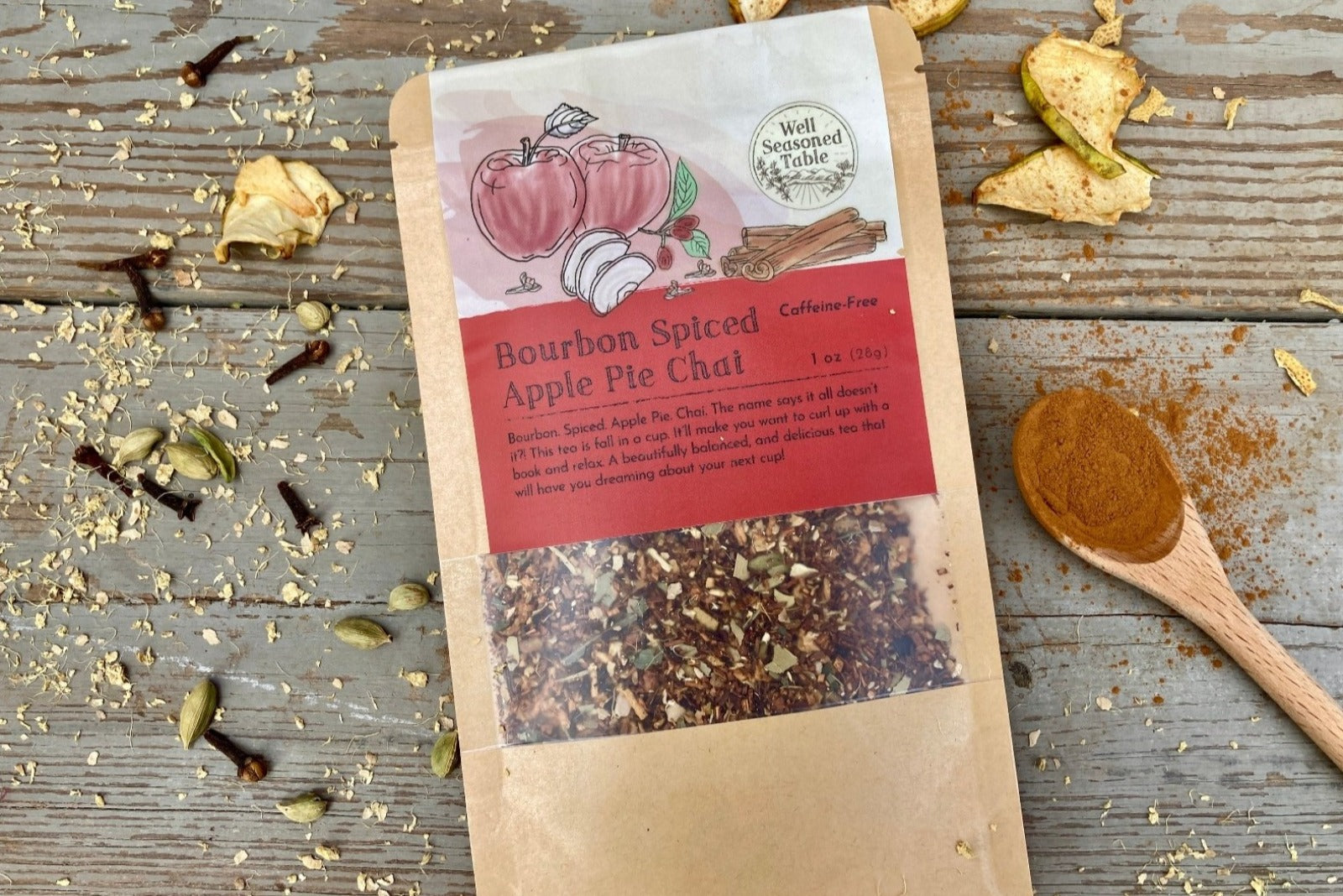 A packet of Bourbon Spiced Apple Pie Chai Tea from Well Seasoned Table on a wooden background with dried apples, spices, and a spoon of cinnamon around it.