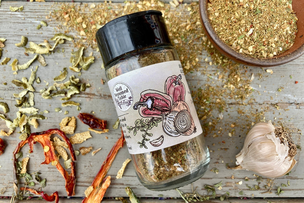 The side of a glass shaker jar of Cuban Seasoning from Well Seasoned Table on a wooden background with dried peppers, garlic, and spices around it.