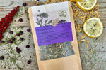 A packet of Elderberry Lemon Tea from Well Seasoned Table on a wooden background with sumac, elderberries, dried lemongrass and lemon slices around it.