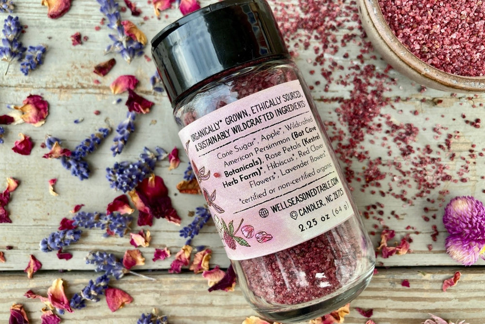 The back of a glass shaker jar of Mountain Flora Sugar from Well Seasoned Table on a wooden background with dried lavender and roses, and a ceramic bowl of bright rose colored sugar.