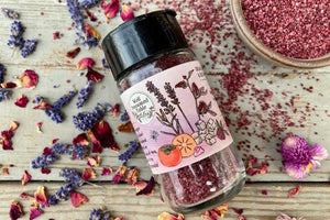 The side of a glass shaker jar of Mountain Flora Sugar from Well Seasoned Table on a wooden background with dried lavender and roses, and a ceramic bowl of bright rose colored sugar.